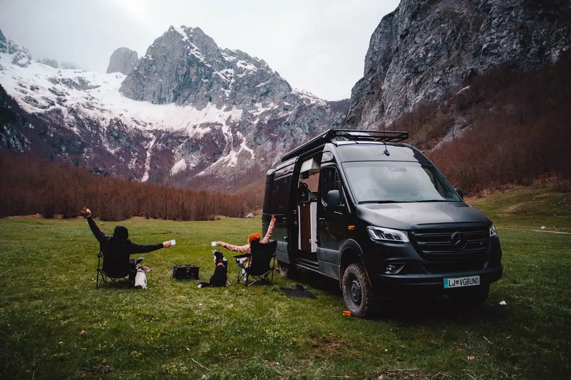 A camper van parked in a snow-capped mountain valley in Montenegro, with travelers relaxing on a road trip